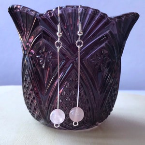 May include: A pair of silver dangle earrings with pink rose quartz beads. The earrings are hanging from a purple glass vase.