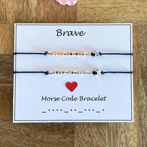 May include: A white card with the word "Brave" printed in black. Two blue strings with silver and rose gold beads and silver star charms are attached to the card. The card also says "Morse Code Bracelet" and shows the Morse code for the word "Brave".