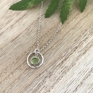 May include: A silver necklace with a delicate chain and a small, round pendant. The pendant features a textured circle with a green gemstone in the center.