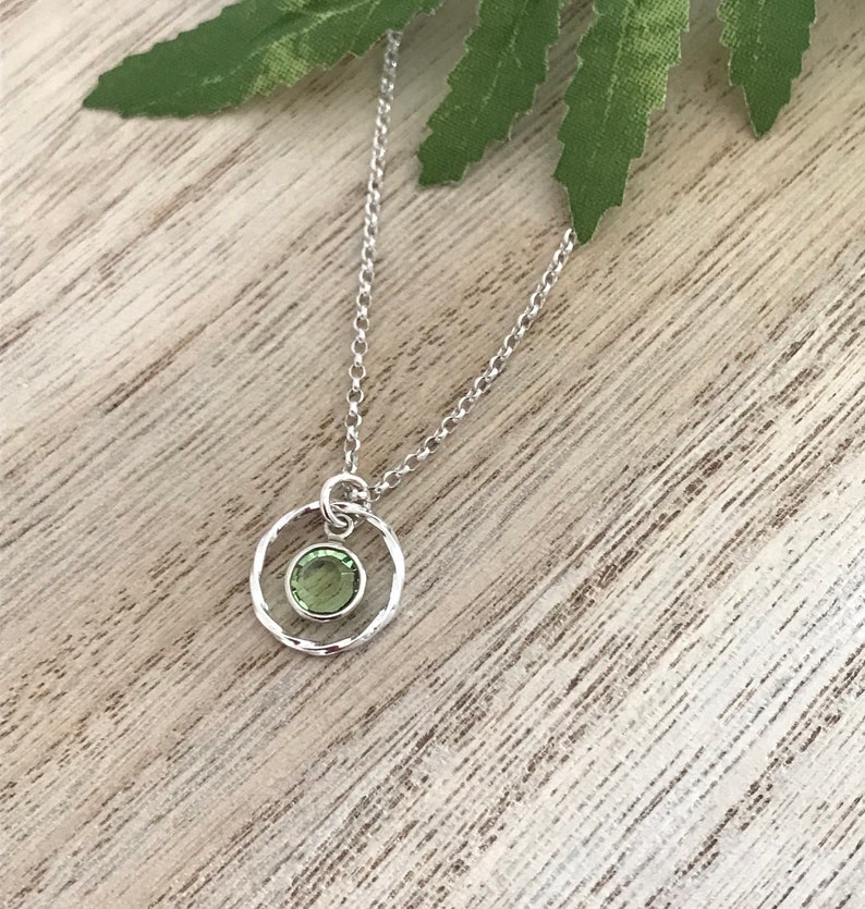May include: A silver necklace with a delicate chain and a small, round pendant. The pendant features a textured circle with a green gemstone in the center.