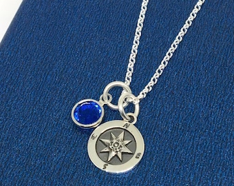 May include: A silver necklace with a compass charm and a blue gemstone. The compass charm has a star design and the letters N, S, E, and W around the edge.