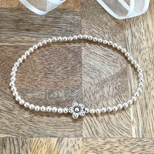 May include: A silver beaded bracelet with a small silver flower charm. The bracelet is on a wooden surface.