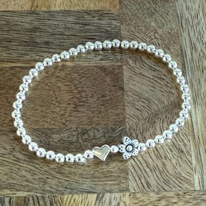 May include: A silver beaded bracelet featuring a heart charm and a flower charm. The bracelet is composed of small, round beads and rests on a wooden surface. The heart charm is plain, and the flower charm has detailed petals.