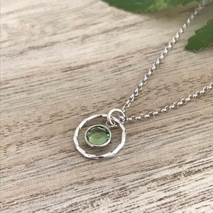 May include: A silver necklace with a delicate chain and a small, hammered circle pendant featuring a green gemstone.