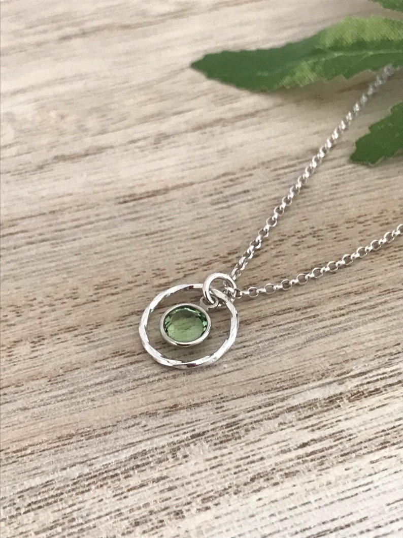 May include: A silver necklace with a delicate chain and a small, hammered circle pendant featuring a green gemstone.