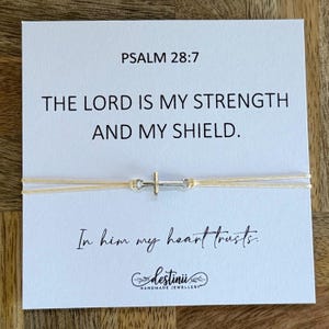 以下が含まれることがあります： 銀色の十字架チャームが付いた繊細なブレスレット。明るい色のコードに通されています。ブレスレットは、「PSALM 28:7」のテキストと「THE LORD IS MY STRENGTH AND MY SHIELD」という引用句が印刷された白いカードの上に置かれています。カードには「In him my heart trusts.」というテキストも含まれています。