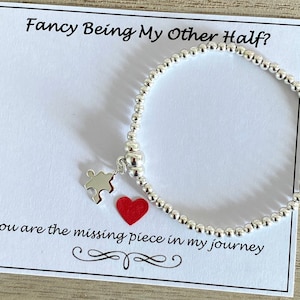 Op de afbeelding: Zilveren armband met een puzzelstukje bedel en een rood hart. De kaart luidt "Fancy Being My Other Half? You are the missing piece in my journey."