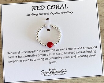 May include: A silver ring with a red coral bead. The ring is on a white card with the text "RED CORAL Sterling Silver & Crystal Jewellery" and a description of the healing properties of red coral.