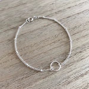 May include: A delicate silver bracelet featuring a chain with small beads and a circular charm. The bracelet is approximately 17.78 cm long and has a lobster clasp closure. The jewelry is displayed on a light brown wooden surface.