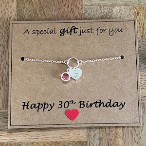 Puede incluir: Un collar de plata con una gema rosa y un dije de corazón grabado con la letra "G". El collar está sobre una tarjeta marrón que dice "A special gift just for you" y "Happy 30th Birthday". Un pequeño corazón rojo está en la parte inferior.