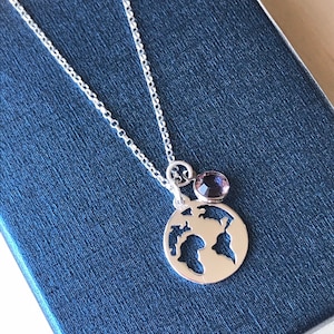 Könnte beinhalten: Eine silberne Halskette mit einer zarten Kette und einem Anhänger, der eine Weltkarte mit einem kleinen violetten Edelstein-Akzent zeigt.
