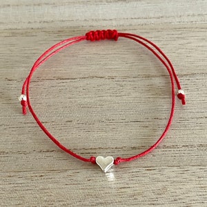 Könnte beinhalten: Ein rotes Armband mit einem silbernen Herz-Anhänger. Das Armband ist oben mit einem Knoten gebunden und hat zwei silberne Perlen auf jeder Seite des Herz-Anhängers.