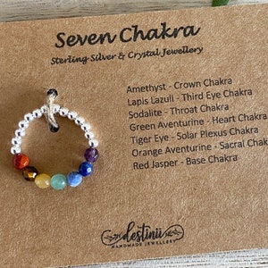Può includere: Un anello in argento sterling con sette pietre dei chakra. Le pietre sono diaspro rosso, occhio di tigre, aventurina verde, sodalite, lapislazzuli, ametista e aventurina arancione. L'anello è su una carta marrone con il testo "Seven Chakra Sterling Silver & Crystal Jewellery" e un elenco delle pietre e dei loro chakra corrispondenti.