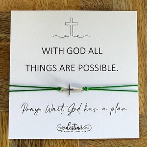 May include: A white card with the text "WITH GOD ALL THINGS ARE POSSIBLE" and "Pray. Wait. God has a plan." A silver cross charm is strung on green cord. The card also has a small cross graphic.