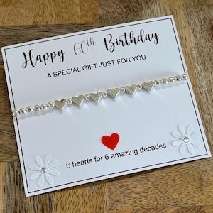 May include: A birthday card with a silver heart bracelet. The card reads "Happy 60th Birthday" and "6 hearts for 6 amazing decades". A red heart and white flower embellishments are also on the card.