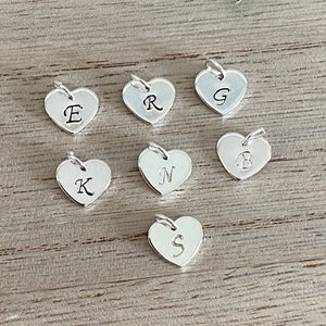 May include: Silver heart-shaped charms, each engraved with a different letter: E, R, G, B, K, N, and S. The charms have a small loop at the top for attaching to a chain or bracelet. The charms are displayed on a wooden surface.