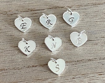 May include: Silver heart-shaped charms, each engraved with a different letter: E, R, G, B, K, N, and S. The charms have a small loop at the top for attaching to a chain or bracelet. The charms are displayed on a wooden surface.