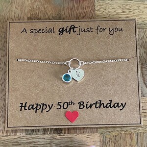 May include: A silver bracelet with a teal-colored gemstone and a heart charm engraved with the letter "H" is displayed on a brown card. The card reads "A special gift just for you" and "Happy 50th Birthday."