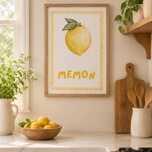 Può includere: Una stampa da parete rettangolare in una cornice di legno chiaro con un'illustrazione ad acquerello di un singolo limone giallo con foglie verdi. La stampa ha un bordo giallo smerlato e la parola MEMON in lettere gialle sotto il limone.