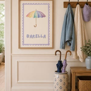 Pode incluir: Uma impressão de parede emoldurada com uma ilustração em aguarela de um guarda-chuva com as cores do arco-íris centrado sobre o texto BARELLA em letras roxas claras, tudo dentro de uma borda festonada roxa clara num fundo creme.