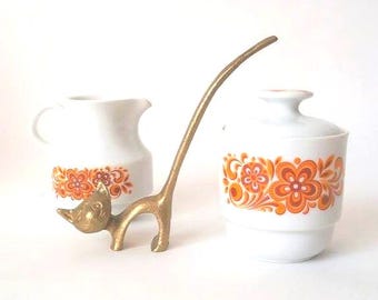 May include: A white ceramic creamer and a lidded sugar bowl, both decorated with a vibrant orange and red floral pattern, positioned alongside a brass cat figurine with an elongated tail that curves upward in a long, slender arc.