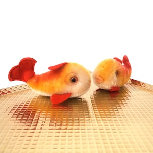 May include: Two plush fish toys with orange and yellow bodies and red fins and tails. The fish have black eyes and are set on a gold, textured surface. One fish is upright, and the other is on its side.