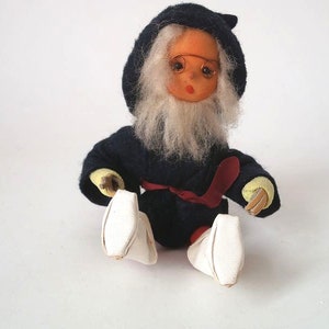 Könnte beinhalten: Eine Vintage-Puppe in einem dunkelblauen Kapuzenmantel mit rotem Schärpe. Die Puppe hat einen weißen Bart, runde Brille und weiße Schuhe. Die Hände der Puppe sind gelb.