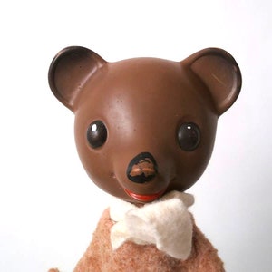 May include: A brown teddy bear with black eyes and a red mouth. The bear is wearing a white scarf and a brown sweater.