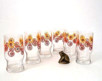 May include: A set of six clear glass tumblers featuring a repeating pattern of stylized sunburst flowers in shades of orange, yellow, and red. A small, dark bronze-colored frog figurine is positioned among the glasses on a plain white surface.