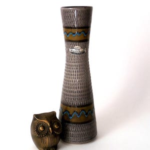 May include: A tall, slender ceramic vase with a textured grey surface, featuring horizontal bands of brown and blue wavy lines, and a small brass owl figurine with detailed feathers and large circular eyes positioned next to it.