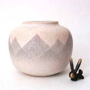 May include: A round ceramic vase with a pale pink base color and a repeating gray triangular mountain pattern around the center. Beside the vase sits a small, dark-colored metal figurine of a rabbit with long ears and a rounded body shape.