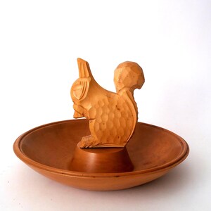 Peut inclure: Un bol en bois brun clair avec une figurine d'écureuil sculptée au centre. Le bol présente un bord circulaire lisse et un bassin peu profond, tandis que la figurine d'écureuil montre des textures sculptées visibles sur le corps et la queue.
