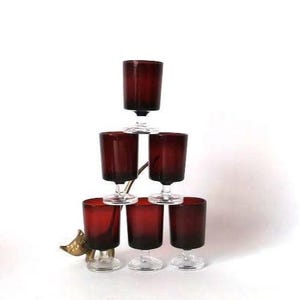 Könnte beinhalten: Ein Set aus sechs tiefroten Glasbechern mit klaren Stielen und Füßen, die in einer Pyramide gestapelt sind. Die Gläser haben eine zylindrische Schalenform und eine glatte Oberfläche. Ein kleiner, metallischer, vogelartiger Dekorationsakzent befindet sich an der Basis.