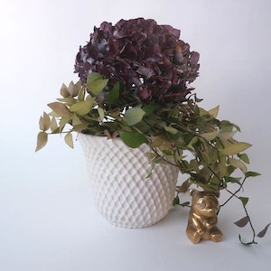 Könnte beinhalten: Ein weißer Keramiktopf mit Diamantmuster, gefüllt mit getrockneten lila Hortensien und grünen Ranken. Eine goldene Bärenfigur steht vor dem Topf.