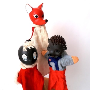 May include: Three hand puppets featuring a red fox with a white scarf, a black-headed character with white hair wearing a red outfit, and a dark brown hedgehog wearing a grey shirt with blue overalls and red trousers, all with distinct facial features.