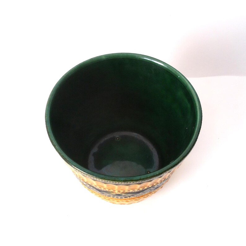 May include: A cylindrical ceramic planter with a deep forest green interior and a glossy finish. The exterior features a textured pattern with alternating bands of yellow and dark green, showcasing a repeating embossed geometric design.