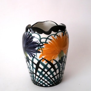 Könnte beinhalten: Eine Keramikvase mit weißem Untergrund und handgemaltem Muster aus dunkelblauen und orangefarbenen Blumen. Das Design umfasst einen schwarzen, gewellten Rand und ein dunkelgrünes Gittermuster, das den unteren Teil der Außenfläche bedeckt.