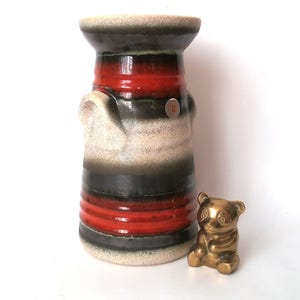 Puede incluir: Un jarrón de cerámica alto con forma cilíndrica, con bandas rojas, negras y beige. Una pequeña figura de panda dorada se encuentra junto al jarrón. El jarrón tiene una superficie texturizada y una pequeña pegatina circular.