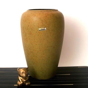 Könnte beinhalten: Eine hohe, olivgrüne Keramikvase mit gesprenkelter Oberfläche. Vor der Vase sitzt eine kleine, goldfarbene Panda-Figur. Die Vase hat einen schwarzen Innenraum und einen kleinen Aufkleber an der Seite.