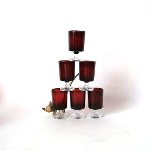 May include: Set of six red glass stemmed shot glasses stacked in a pyramid shape. The glasses are clear with a red tint and have a classic design.