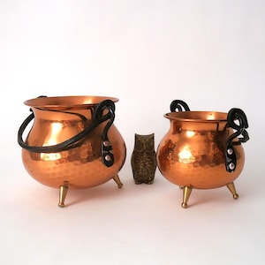 May include: Two copper cauldrons with black handles and brass-colored feet. The cauldrons have a hammered texture and are displayed with a small bronze owl figurine. The cauldrons are a decorative item.