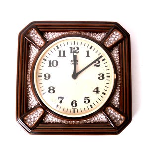 Könnte beinhalten: Quadratische Wanduhr mit braunem Rahmen und weißem Zifferblatt. Das Zifferblatt hat schwarze Ziffern und Zeiger, die Uhrzeit zeigt kurz nach 1:00 Uhr an. Der Rahmen hat ein gewebtes Design mit einem gesprenkelten Muster.