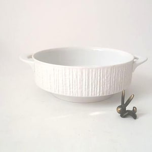 Könnte beinhalten: Weißer Keramiknapf mit strukturierten vertikalen Linien und zwei Griffen. Neben der Schüssel befindet sich eine kleine, bronzefarbene Hasenfigur. Die Schüssel hat ein breites, flaches Design.