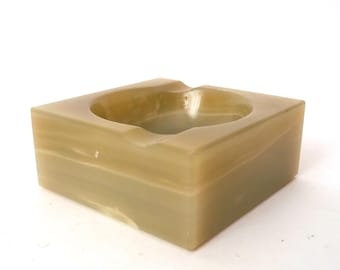 Pode incluir: Um cinzeiro quadrado feito de pedra verde-claro e bege. Possui uma reentrância circular no centro e uma ranhura para cigarros. O cinzeiro é um item decorativo, medindo aproximadamente 10 cm x 10 cm.