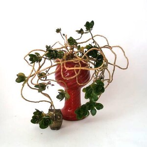 Könnte beinhalten: Ein leuchtend roter Keramik-Pflanztopf mit einem Wirbelmuster, der eine Sukkulentenpflanze mit grünen Blättern und hellbraunen Stängeln enthält. Eine kleine Eulenfigur aus Bronze steht neben dem Topf.