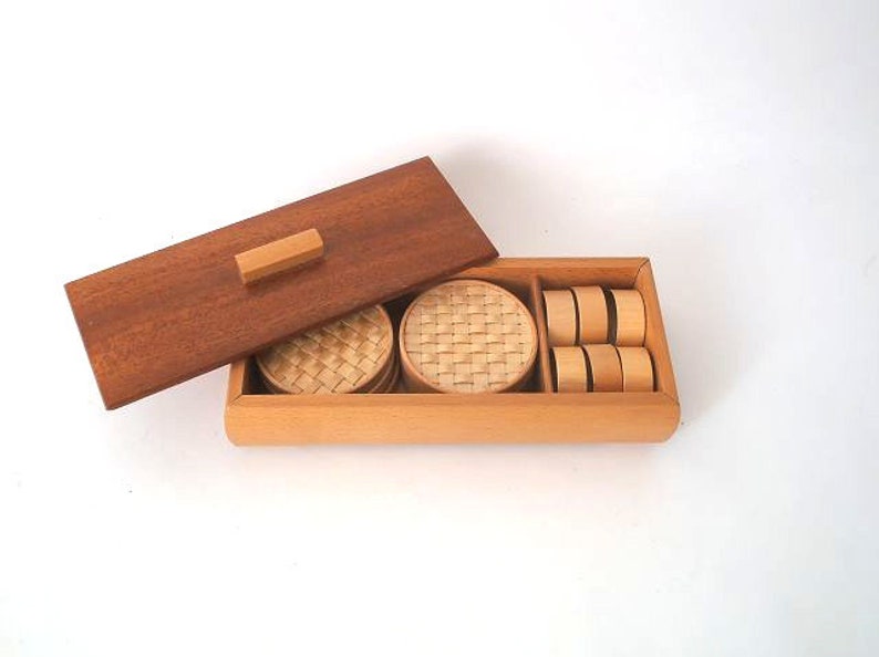May include: A wooden box with a lid, containing woven coasters and round wooden coasters. The box is made of light wood and has a dark wood lid.