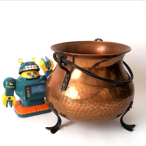 May include: A hammered copper cauldron with a wide rim, a black metal swing handle, and three curved black metal legs, positioned next to a teal, yellow, and orange plastic robot toy with a screen on its chest and raised arms.