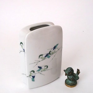 May include: A white ceramic vase with a rectangular shape and a curved top. The vase is decorated with painted birds on branches in shades of blue, green, and white. A small bronze bird figurine sits beside the vase.