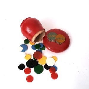 May include: A red wooden container with a matching lid featuring the text FLOH SPIEL FUR 6 SPIELER, accompanied by a collection of small, flat, circular game pieces in various colors including red, yellow, green, blue, black, and white.
