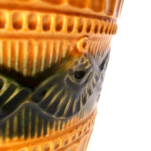 May include: A glazed ceramic vessel featuring a warm amber-orange base color with a prominent dark green, embossed wave-like pattern encircling the exterior. The rim displays a textured, repeating vertical notch design.